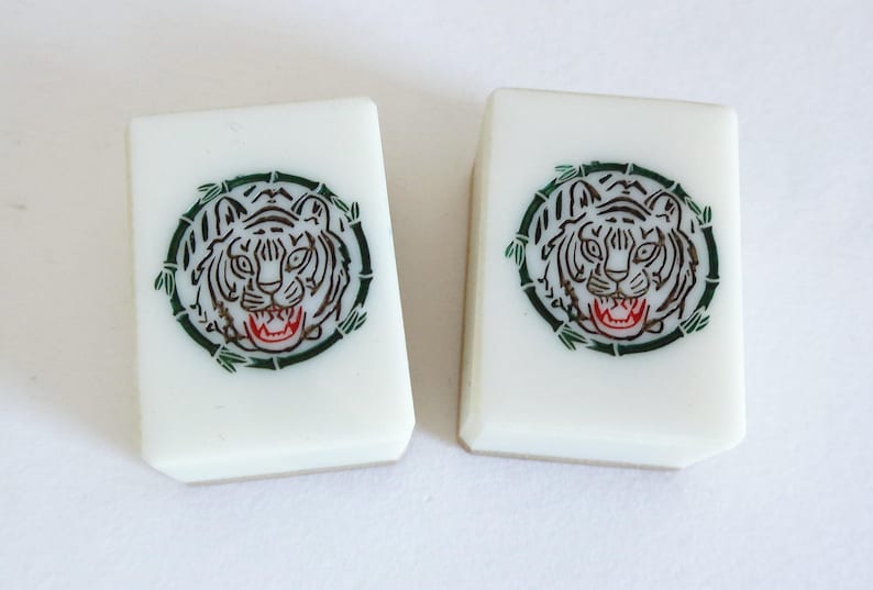 May include: Two white Mahjong tiles with a tiger face design. The tiger is encircled by a green bamboo-like border. The tiger's face is black and white with a red mouth.
