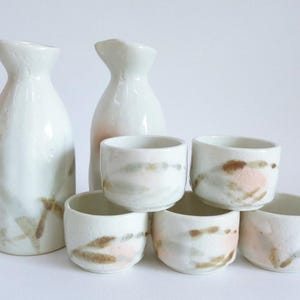 Puede incluir: Conjunto de siete tazas y botellas de sake de cerámica blanca con diseños abstractos marrones y rosas. Las tazas son más pequeñas y las botellas más grandes.