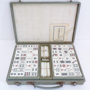 May include: An open vintage Mahjong set in a grey case. The set contains ivory-coloured tiles with red and black markings, dice, and a booklet with Japanese text. The case is open, displaying the organised arrangement of the tiles.