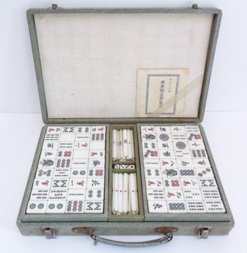 May include: An open vintage Mahjong set in a grey case. The set contains ivory-coloured tiles with red and black markings, dice, and a booklet with Japanese text. The case is open, displaying the organised arrangement of the tiles.