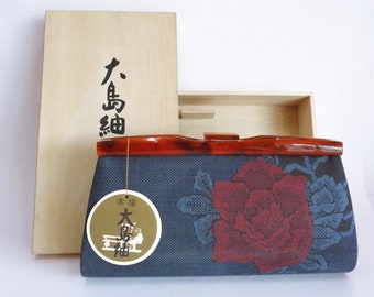 May include: A blue and red floral clutch purse with a brown plastic handle. The purse is in a wooden box with the Japanese characters "大島紬" (Oshima Tsumugi) and "六島紬" (Rokushima Tsumugi) on the box.