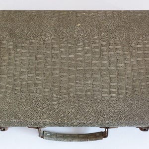 May include: A vintage briefcase with a textured, crocodile-like pattern in shades of gray and green. The briefcase has a handle and metal clasps. The overall design suggests a classic, possibly retro, aesthetic.