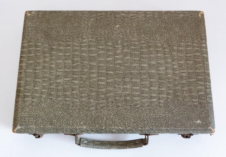 May include: A vintage briefcase with a textured, crocodile-like pattern in shades of gray and green. The briefcase has a handle and metal clasps. The overall design suggests a classic, possibly retro, aesthetic.