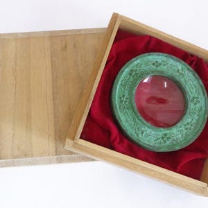 Op de afbeelding: Een ronde, groene bronzen schotel met een decoratief bloemmotief rond de rand. De schotel bevindt zich in een houten doos bekleed met rood fluweel.