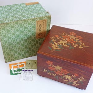 May include: A square wooden box with a dark brown finish, featuring painted orange and yellow floral designs on the lid and front. Beside it sits a green patterned storage box with Japanese text and two small rectangular paper inserts with text.