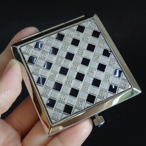May include: A silver compact mirror with a geometric design. The square face features a checkerboard pattern of black and white squares, with small, sparkling accents. The edges are silver, and a small blue jewel is visible on the side.