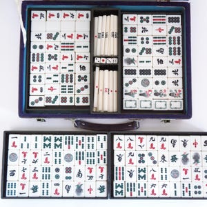 May include: A complete Mahjong set in a blue case, with tiles arranged in trays. The tiles are white with black, red, and green symbols. Dice and tile racks are also included. The set is open, displaying all the components.