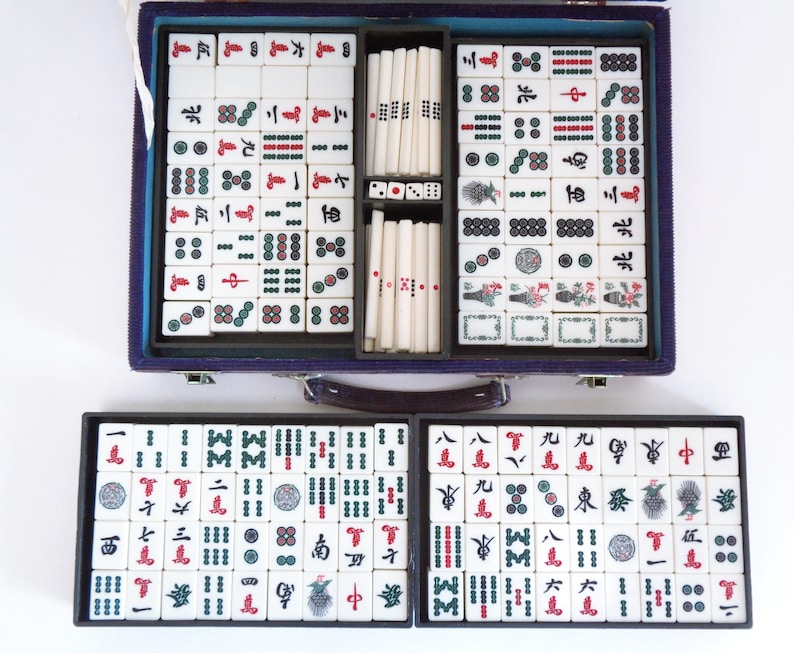 May include: A complete Mahjong set in a blue case, with tiles arranged in trays. The tiles are white with black, red, and green symbols. Dice and tile racks are also included. The set is open, displaying all the components.