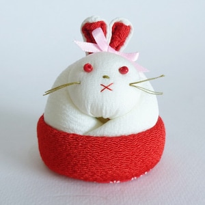 May include: A white fabric rabbit with red ears and a pink bow. The rabbit is sitting on a red fabric base.