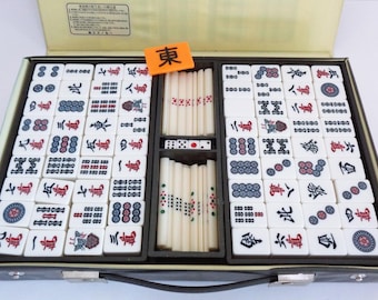 May include: A black carrying case with a set of white mahjong tiles. The tiles have red and blue characters and symbols. The case has a handle and a compartment for the tiles. The case also has a compartment for mahjong sticks and dice.