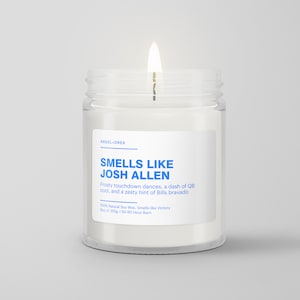 May include: A clear glass jar candle with a white label that reads "Smells Like Josh Allen" and "Frosty touchdown dances, a dash of QB cool, and a zesty hint of Bills bravado". The label also says "100% Natural Soy Wax, Smells like Victory" and "8oz. e 255g. 150-60 Hour Burn".
