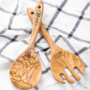 Può includere: Due posate da insalata in legno, un cucchiaio e una forchetta, con finitura marrone chiaro e motivo a venature naturali. Il cucchiaio ha una testa arrotondata e la forchetta ha tre rebbi. Il testo "Michelle's Kitchen" è inciso sul manico.