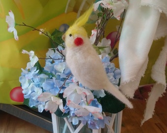 Könnte beinhalten: Ein weißer und gelber Filzvogel, der auf einem Zweig mit blauen und weißen Blüten sitzt. Der Vogel sitzt in einer weißen Laterne mit einem Glasfenster.