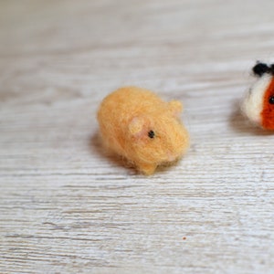 May include: Two small, hand-felted guinea pigs. One is orange and the other is white with orange accents. They are both sitting on a white wooden surface.
