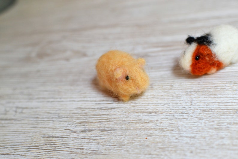 May include: Two small, hand-felted guinea pigs. One is orange and the other is white with orange accents. They are both sitting on a white wooden surface.