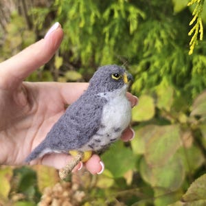 May include: A handmade felt bird figurine in shades of gray and white, perched on a small twig. The bird has a yellow beak and is held in a hand against a backdrop of green foliage. The craft bird is a unique decorative item.