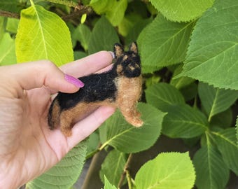 May include: A miniature felted German Shepherd dog figurine, featuring black and tan coloring. The handcrafted dog is held against a backdrop of green leaves. The figurine is a detailed representation of the breed.