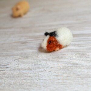 May include: A white and brown felt guinea pig with black eyes and a black felt bow on its head.