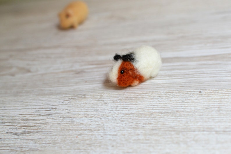 May include: A white and brown felt guinea pig with black eyes and a black felt bow on its head.
