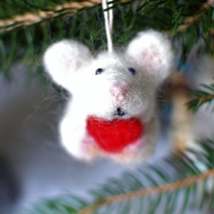 May include: A handmade white felt mouse Christmas ornament with a red heart-shaped chest. The mouse has black bead eyes, a pink nose, and a white string for hanging. The ornament is hanging on a green pine tree branch.