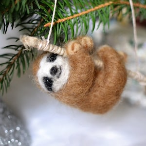 May include: A brown and white felt sloth ornament hanging from a branch. The sloth has a cute face with black eyes and a white nose.