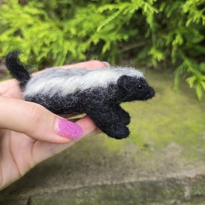 以下が含まれることがあります： 手作りのフェルト製のイタチの置物。イタチは黒い体と背中の白い縞模様があります。小さな動物は黒い頭と尾を持っています。置物は、緑の葉を背景に手で持たれています。