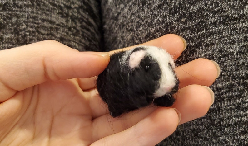 May include: A small, black and white needle felted guinea pig. The guinea pig has pink ears and black eyes.