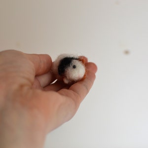 May include: A small, hand-felted guinea pig figurine with white, black, and brown wool. The figurine is being held in a person's hand.