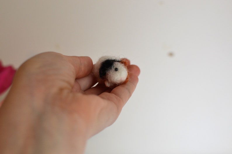 May include: A small, hand-felted guinea pig figurine with white, black, and brown wool. The figurine is being held in a person's hand.
