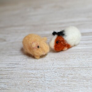 May include: Two needle felted guinea pigs. One is brown and white with black eyes and a black nose. The other is light brown with black eyes and a pink nose. They are both sitting on a light wood surface.