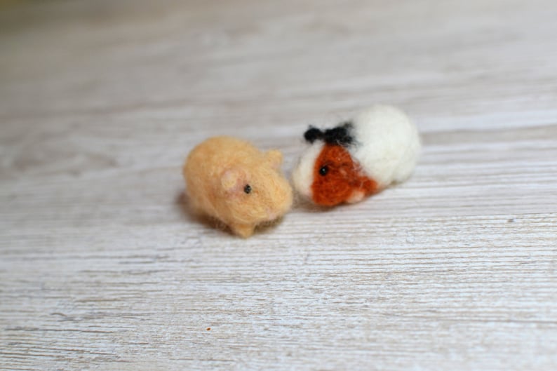 May include: Two needle felted guinea pigs. One is brown and white with black eyes and a black nose. The other is light brown with black eyes and a pink nose. They are both sitting on a light wood surface.