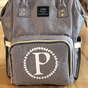 May include: Gray diaper bag backpack with gold zipper accents and a white monogram of the letter "P" in a wreath design. The bag has a label that reads "LAND MOMMYSEARLYEAS CHIC MOM GIVE YOUTHE BESTCARE".