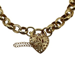 Op de afbeelding: Een goudkleurige kettingarmband met een hartvormige hanger. De hanger is ingewikkeld ontworpen met bloempatronen en een kleine ketting. De armband heeft grote, afgeronde schakels.