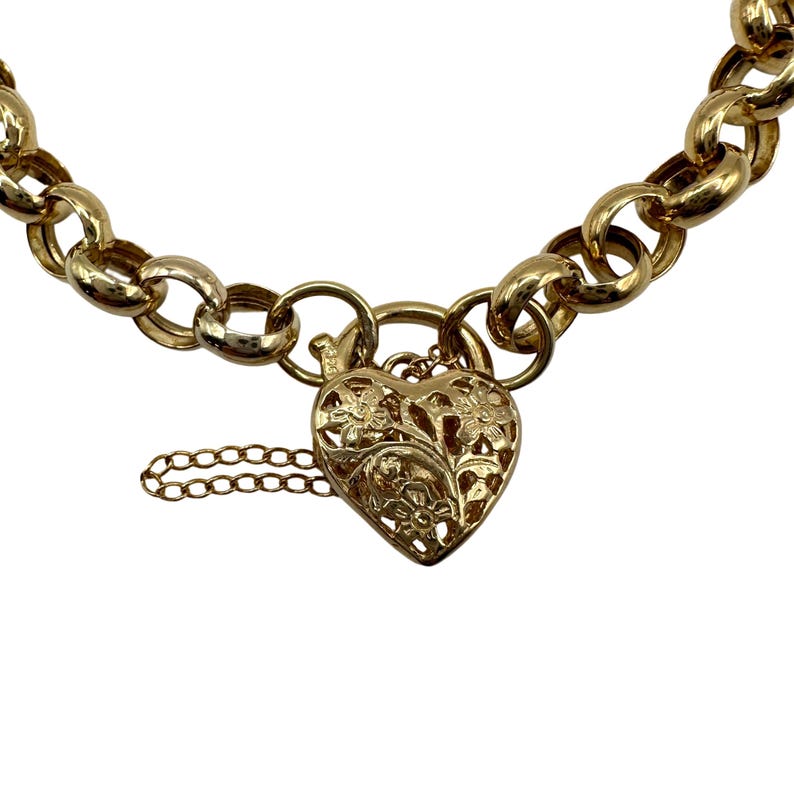 Op de afbeelding: Een goudkleurige kettingarmband met een hartvormige hanger. De hanger is ingewikkeld ontworpen met bloempatronen en een kleine ketting. De armband heeft grote, afgeronde schakels.