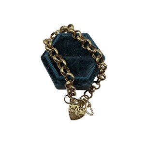 Op de afbeelding: Een goudkleurige armband met een schakelkettingontwerp en een hartvormige bedel. De bedel heeft een gegraveerd bloemmotief en een kleine ketting. De armband wordt gepresenteerd op een donkerblauwe fluwelen ondergrond.