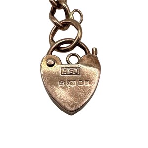 May include: A heart-shaped gold padlock with a chain. The padlock has the initials "ASJ" and the number "375" engraved on it. The chain is made of gold links. The padlock and chain are set against a white background.