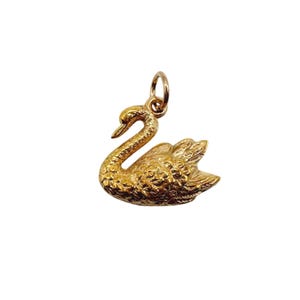 May include: A gold-toned metal pendant shaped like a swan, featuring detailed textured feather patterns across its body and wings. The swan is depicted with its neck curved gracefully, and a small circular jump ring is attached to the top.