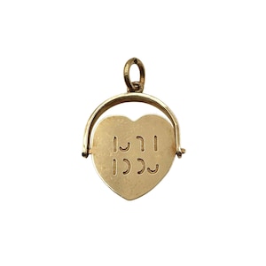May include: A gold-toned heart-shaped pendant featuring a swivel mechanism held by a curved metal frame. The center of the heart displays embossed, stylized lettering that appears to be a sequence of vertical lines and curved marks.