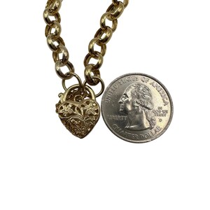 Op de afbeelding: Een goudkleurige ketting met een hartvormig medaillon. Het medaillon heeft een bloemenpatroon en staat naast een Amerikaanse kwartdollar ter vergelijking. De ketting heeft een ketting van grote, ronde schakels.