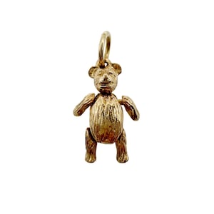 May include: A gold-toned metal charm in the shape of a teddy bear with a textured surface finish. The bear has a rounded head, small ears, and articulated limbs. A circular jump ring is attached to the top of the bear's head for hanging purposes.