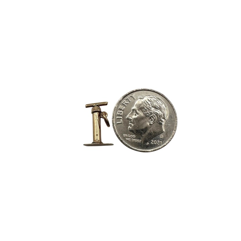 May include: A small gold-toned metal charm shaped like a manual air pump, featuring a T-shaped handle and a circular base. The charm is positioned next to a standard United States dime for scale, which measures approximately 0.705 inches in diameter.