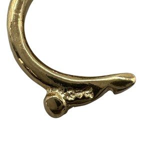 Op de afbeelding: Close-up van een goudkleurige metalen oorbel. De oorbel heeft een gebogen ontwerp met een kleine, ronde opening en een getextureerd oppervlak. Het metaal heeft een glanzende afwerking.