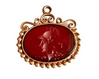 May include: A gold pendant with a red cameo featuring a profile of a woman wearing a helmet. The cameo is set in a gold frame with a beaded border.