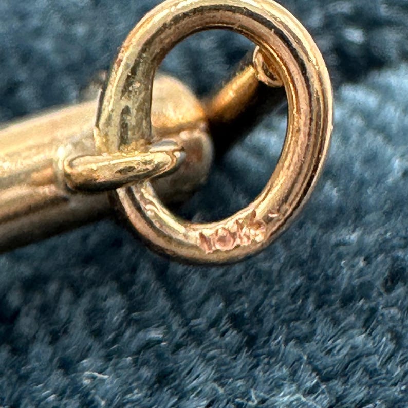 May include: A gold-toned metal jump ring attached to a cylindrical metal component. The surface of the circular jump ring features a small, stamped inscription that reads 10K, indicating the metal purity of the jewelry finding.