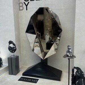May include: A large, faceted, silver mirror with a black base, standing on a concrete floor. The mirror is shaped like a diamond and reflects the surrounding space. The text "PUBLISHED BY" is written in black letters on a white wall behind the mirror. The text "BURBERRY" is written in black letters on the floor in front of the mirror.