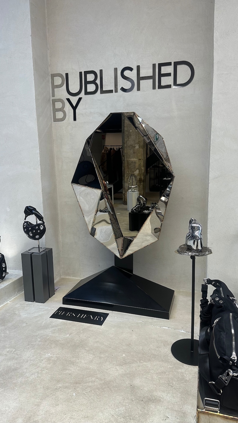 May include: A large, faceted, silver mirror with a black base, standing on a concrete floor. The mirror is shaped like a diamond and reflects the surrounding space. The text "PUBLISHED BY" is written in black letters on a white wall behind the mirror. The text "BURBERRY" is written in black letters on the floor in front of the mirror.