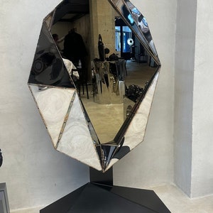May include: A large, faceted, silver mirror with a black base. The mirror is shaped like a diamond and is standing on a black pedestal. The base has the text "PIERS HENRY" printed on it.