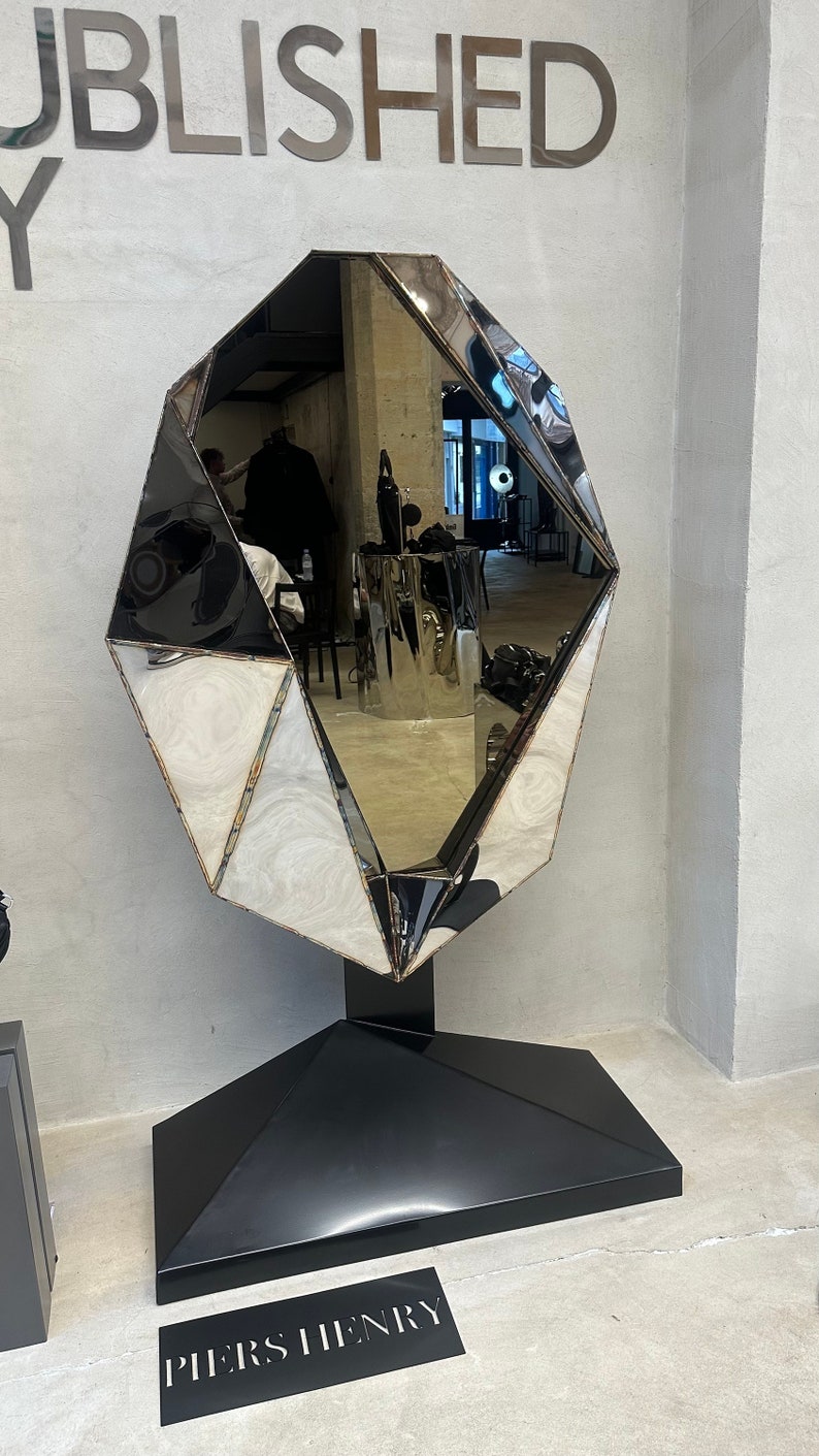 May include: A large, faceted, silver mirror with a black base. The mirror is shaped like a diamond and is standing on a black pedestal. The base has the text "PIERS HENRY" printed on it.
