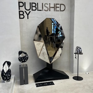 May include: A black and silver geometric mirror with a black base and the text "PUBLISHED BY" on the wall behind it. Three black bags with silver studs are displayed on stands in front of the mirror.