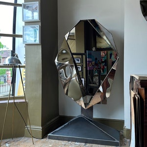 May include: A large, faceted, silver mirror with a geometric design. The mirror is standing on a black base in a room with wooden floors.